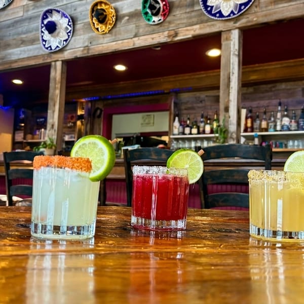 Three colorful margaritas with lime garnishes on a rustic wooden table in a vibrant Mexican restaurant.