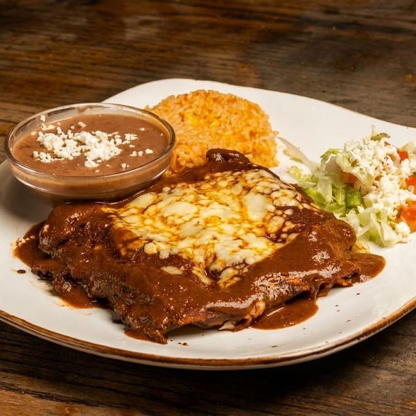 A plate of enchiladas covered in mole sauce and melted cheese, with rice, refried beans, and salad.