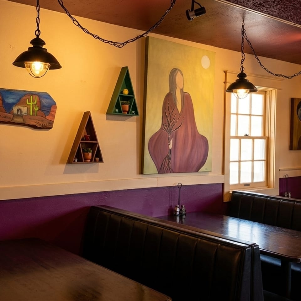 Restaurant interior with booth seating, hanging lights, and colorful artwork on cream-colored walls.